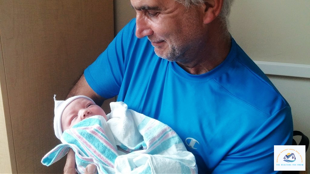 A smiling grandfather in a bright blue shirt holds his newborn grandchild, wrapped in a hospital blanket. This heartwarming moment reflects the joy of family and homeownership, making Bloomfield Hills, MI, a great place to settle down. Trusted realtors in Bloomfield Hills help families find the perfect home for creating lasting memories…