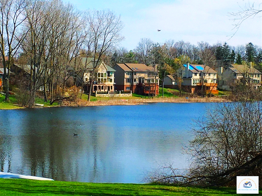 A stunning view of lakefront homes for sale in West Bloomfield, MI, with serene waters reflecting the picturesque houses and surrounding trees. These waterfront properties offer private decks, peaceful nature views, and direct access to the lake…