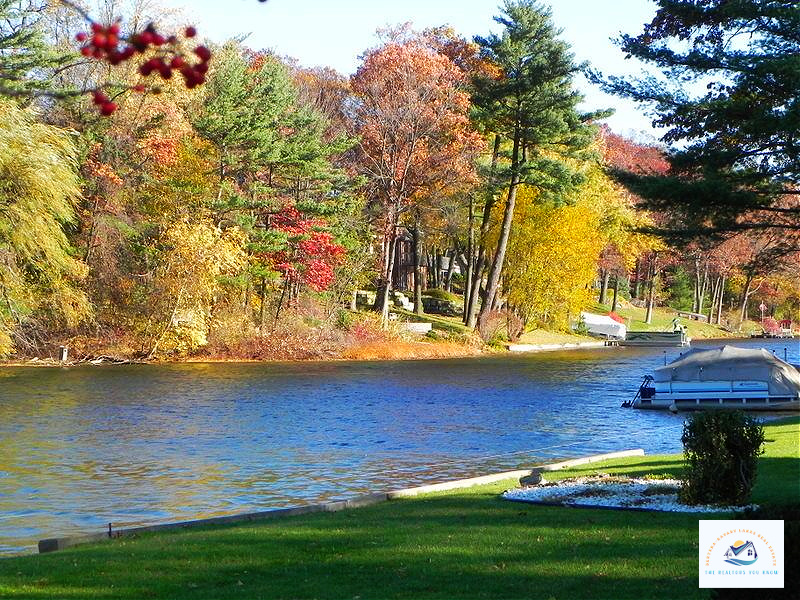 A vibrant autumn lakeside view in Oakland County, MI, featuring private docks, colorful trees, and a pontoon boat. This picturesque setting highlights the beauty of waterfront living in Michigan…