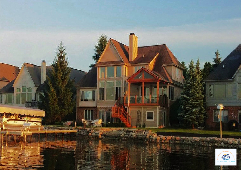 A breathtaking lakefront home in West Bloomfield, MI, illuminated by the golden hues of sunset. This elegant property features large glass windows, a covered balcony, and a charming wooden staircase leading to the landscaped backyard by the water. The home offers stunning lake views and private dock access, making it an excellent choice for those seeking lakefront homes for sale in West Bloomfield, MI…