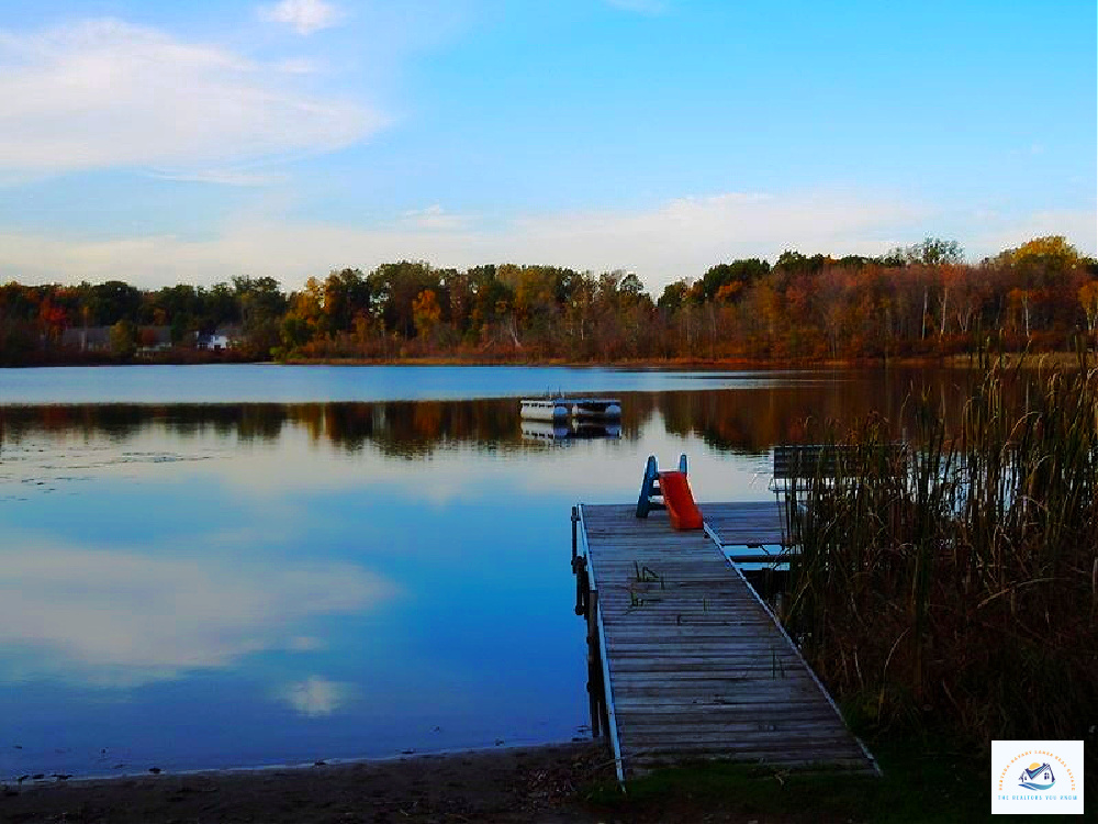A peaceful lakefront scene in West Bloomfield, MI, featuring a private wooden dock with a slide leading into the calm water. The tranquil lake reflects the blue sky and surrounding trees, making it an ideal setting for waterfront living…