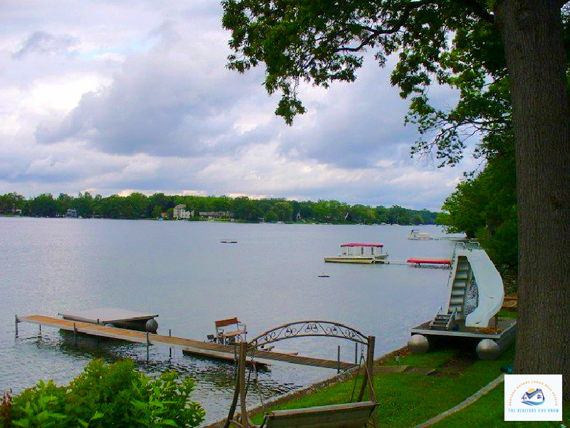 A picturesque view of a private dock and waterfront seating area overlooking a serene lake in West Bloomfield, MI. The image captures a well-maintained backyard with a lakeside swing, a boat dock, and a waterslide, making it a perfect setting for enjoying Michigan lakefront living…