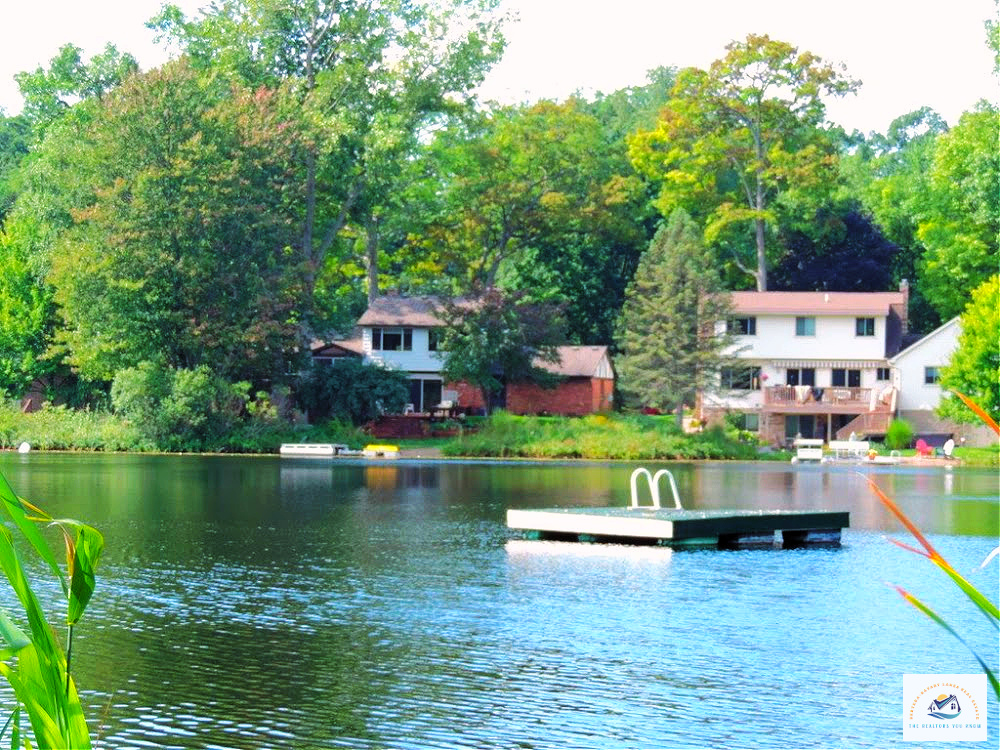 A beautiful view of West Bloomfield, MI waterfront homes with lush greenery surrounding the lake. A floating swim platform sits on the calm water, making this lakefront property ideal for relaxation and outdoor enjoyment…