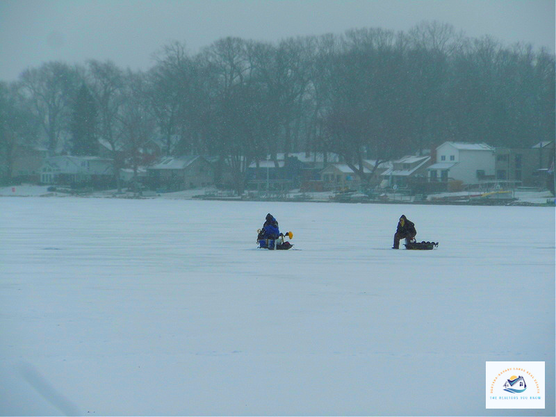 Two people ice fishing on a frozen lake in West Bloomfield, MI, with waterfront homes visible in the distance. This area offers year-round lake activities and is a great spot for buyers looking for Michigan lakefront homes…